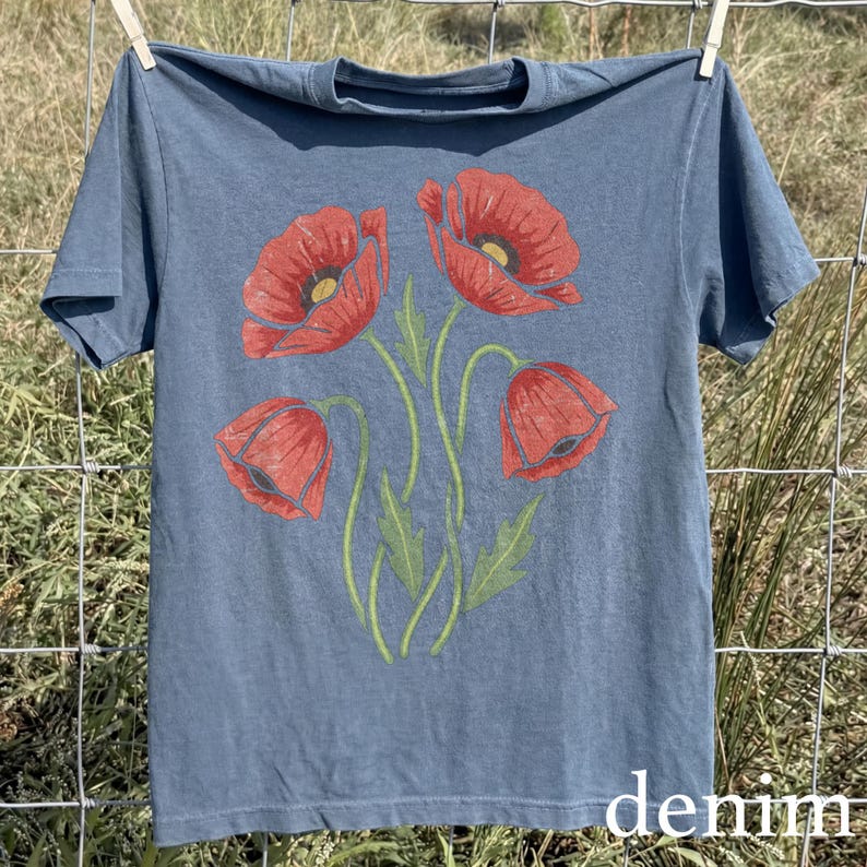 Op de afbeelding: Een denim blauw t-shirt met een bloemenontwerp van rode klaprozen met groene stelen en bladeren. Het woord "denim" staat in het wit rechtsonder gedrukt. Het shirt hangt aan een waslijn.