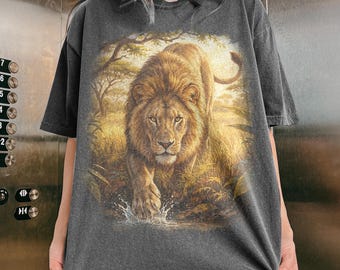 May include: Dark grey t-shirt with a detailed illustration of a lion walking through water. The lion has a golden mane and is set against a backdrop of savanna vegetation. The word "pepper" is printed at the bottom.