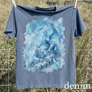 May include: Denim blue t-shirt featuring a detailed illustration of a blue dragon perched on a rocky, icy landscape. The dragon has large wings and is set against a snowy background. The word "denim" is printed at the bottom.