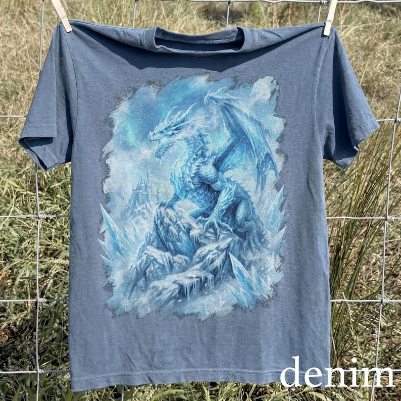 May include: Denim blue t-shirt featuring a detailed illustration of a blue dragon perched on a rocky, icy landscape. The dragon has large wings and is set against a snowy background. The word "denim" is printed at the bottom.
