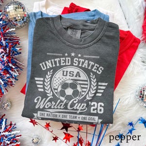 May include: A charcoal grey short-sleeve t-shirt featuring a white graphic print that reads United States USA World Cup '26 One Nation One Team One Goal with a soccer ball and flag motif. The shirt has a crew neckline and a Comfort Colors brand label.