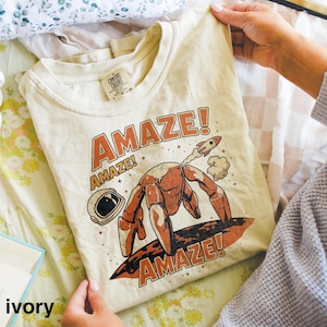 May include: Ivory-colored t-shirt with a crew neck. The shirt features a graphic of a robot-like figure surfing on a board, with a rocket ship and the word "AMAZE!" in orange. The shirt is held by a person.