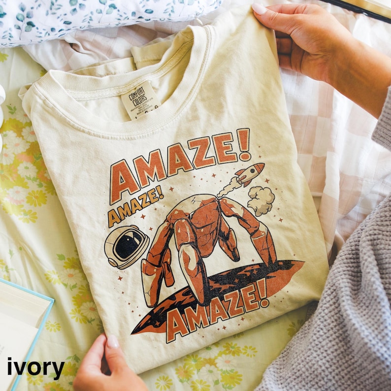 May include: Ivory-colored t-shirt with a crew neck. The shirt features a graphic of a robot-like figure surfing on a board, with a rocket ship and the word "AMAZE!" in orange. The shirt is held by a person.