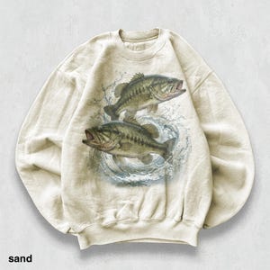 May include: Sand-coloured sweatshirt with a detailed illustration of two largemouth bass leaping from water. The fish are depicted in shades of green, brown, and grey, with open mouths and water splashing around them. The word "sand" is printed at the bottom.
