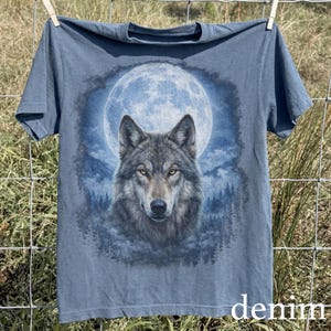 May include: Denim blue t-shirt featuring a detailed wolf portrait set against a full moon and forest backdrop. The wolf has yellow eyes and gray fur. The word "denim" is printed in white at the bottom of the shirt.