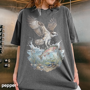 May include: Dark grey t-shirt featuring a detailed graphic of an osprey catching a fish in a lake. The design includes water splashes, aquatic plants, and a colorful fish. The word "pepper" is printed at the bottom left.