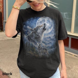 May include: Black t-shirt featuring a detailed graphic of two wolves howling at a full moon. The design includes a forest and mountain landscape in shades of blue and gray. The word "black" is printed at the bottom.