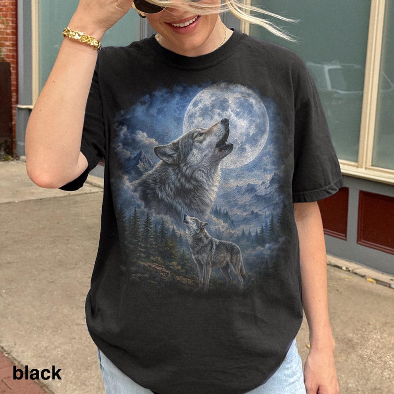 May include: Black t-shirt featuring a detailed graphic of two wolves howling at a full moon. The design includes a forest and mountain landscape in shades of blue and gray. The word "black" is printed at the bottom.