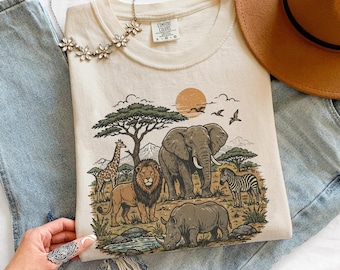 May include: A cream-colored t-shirt featuring a graphic print of an elephant, lion, giraffe, zebra, and rhinoceros in a savanna landscape with a sun and trees. The shirt has a crew neckline and a Comfort Colors brand label on the inner collar.