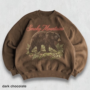 May include: Dark chocolate-colored sweatshirt featuring a graphic of a bear walking through ferns, with the words "Smoky Mountains" in red script above. The text "dark chocolate" is at the bottom of the image.