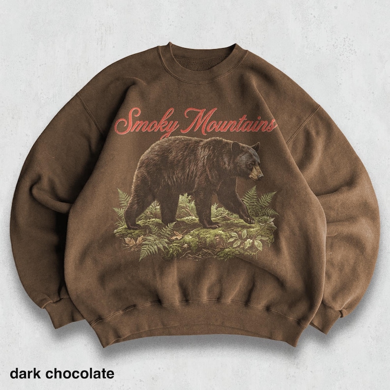 May include: Dark chocolate-colored sweatshirt featuring a graphic of a bear walking through ferns, with the words "Smoky Mountains" in red script above. The text "dark chocolate" is at the bottom of the image.