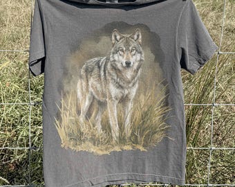 May include: A grey t-shirt with a detailed illustration of a wolf standing in tall grass. The wolf is depicted in shades of grey, brown, and white, with a focused gaze. The word "pepper" is printed at the bottom of the shirt.