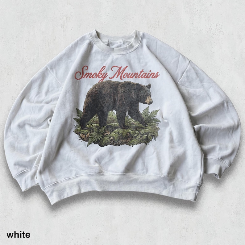 May include: White sweatshirt featuring a graphic of a black bear walking on a bed of ferns, with "Smoky Mountains" in red script above. The word "white" is printed at the bottom. A casual, comfortable top.