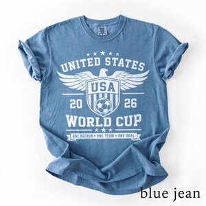 May include: A blue short-sleeved t-shirt featuring a white graphic print that reads UNITED STATES, USA, 2026, WORLD CUP, and ONE NATION ONE TEAM ONE GOAL. The design includes a central shield with a soccer ball, an eagle, and stars.