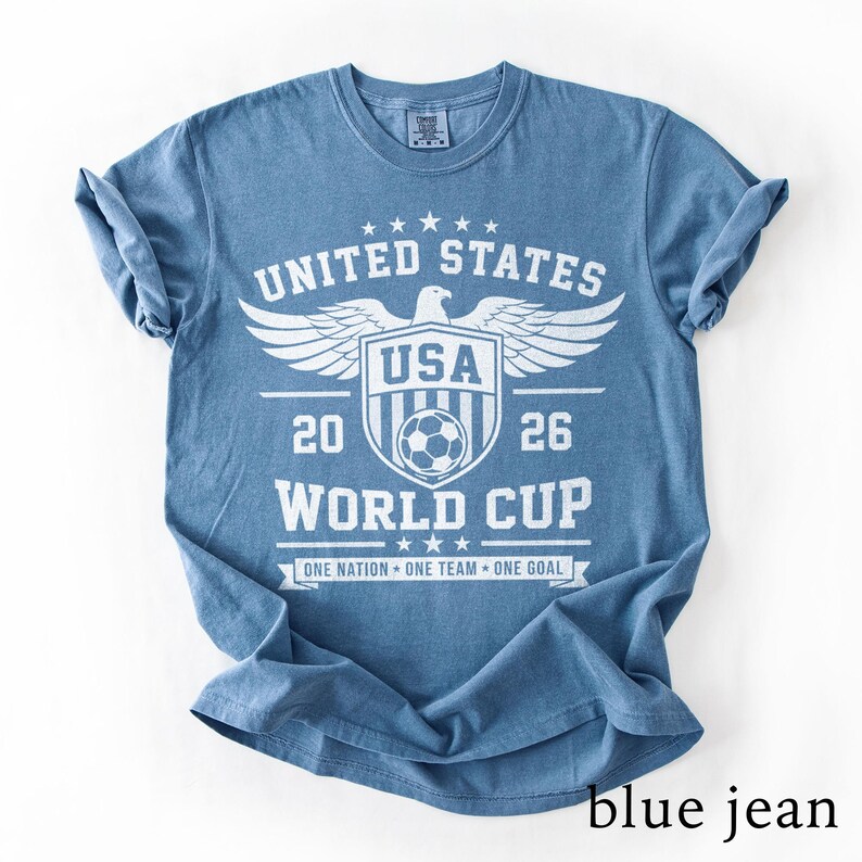 May include: A blue short-sleeved t-shirt featuring a white graphic print that reads UNITED STATES, USA, 2026, WORLD CUP, and ONE NATION ONE TEAM ONE GOAL. The design includes a central shield with a soccer ball, an eagle, and stars.