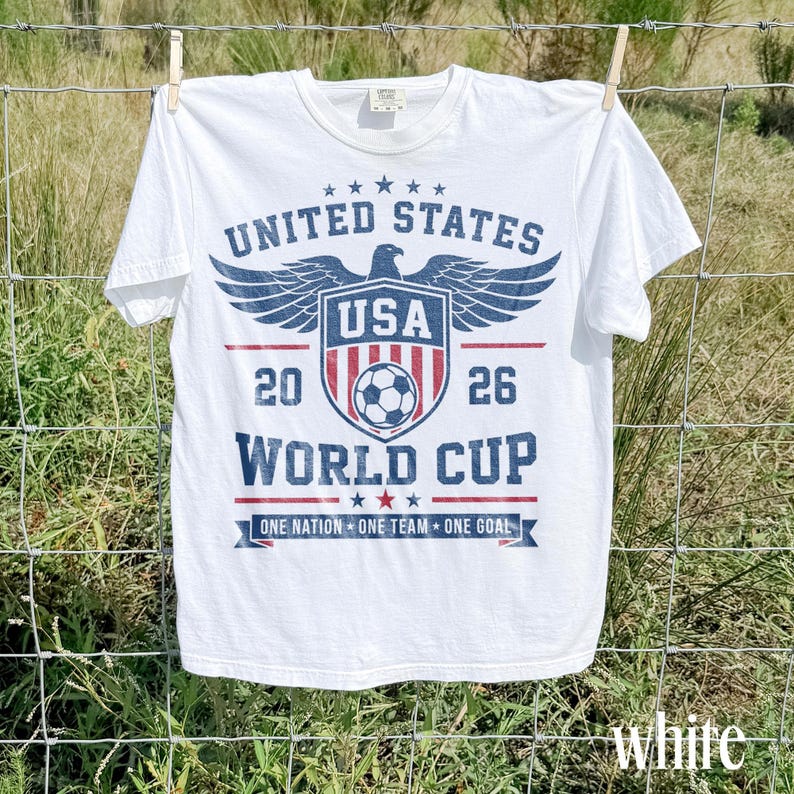 May include: A white short-sleeve t-shirt featuring a blue and red graphic with an eagle, a shield, a soccer ball, and the text UNITED STATES 2026 WORLD CUP ONE NATION ONE TEAM ONE GOAL. The design has a distressed, vintage-inspired appearance.