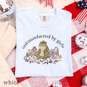 May include: A light blue Comfort Colors sweatshirt with the phrase "outnumbered by girls" in a curved font. The graphic features a frog wearing a cowboy hat surrounded by makeup and toys. The word "white" is at the bottom.