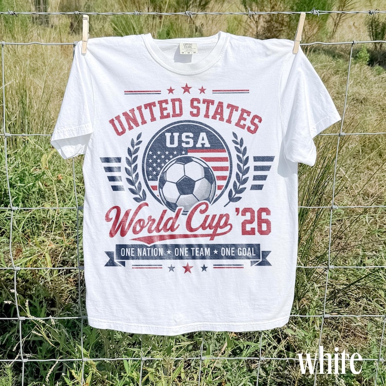 May include: A white short-sleeve t-shirt featuring a red, white, and blue graphic print. The design includes a soccer ball, the text UNITED STATES, USA, World Cup '26, and the phrase ONE NATION ONE TEAM ONE GOAL with stars and laurel accents.