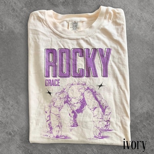 May include: Ivory-colored t-shirt with the word "ROCKY" in a purple, outlined font, and the word "GRACE" below it. A purple illustration of a rock formation is centered below the text. The shirt is folded.