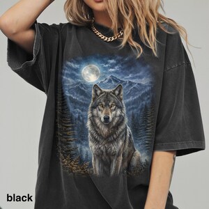 May include: Black oversized t-shirt featuring a detailed graphic of a wolf under a full moon in a forest setting. The wolf has gray and brown fur, and the background includes mountains and pine trees. The word "black" is printed at the bottom.
