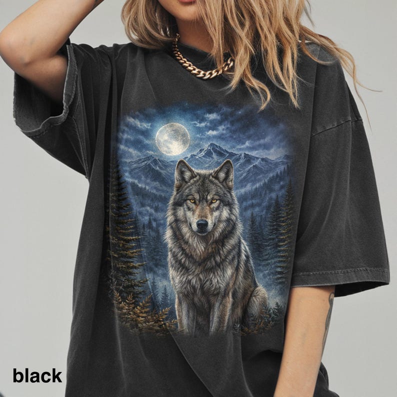 May include: Black oversized t-shirt featuring a detailed graphic of a wolf under a full moon in a forest setting. The wolf has gray and brown fur, and the background includes mountains and pine trees. The word "black" is printed at the bottom.