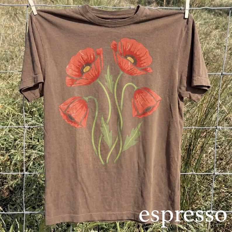 Op de afbeelding: Espresso-kleurig t-shirt met een bloemenontwerp van vier rode klaprozen met groene stelen en bladeren. Het shirt hangt aan een waslijn. Het woord "espresso" staat onderaan gedrukt.