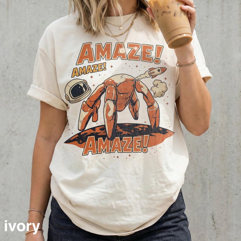 May include: Ivory-colored t-shirt with a retro graphic design. The design features a crab-like creature with a rocket, astronaut helmet, and the word "AMAZE!" in orange. The shirt has short sleeves and a relaxed fit.