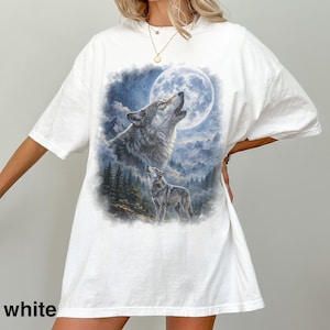 May include: White t-shirt featuring a detailed graphic of two wolves howling at a full moon. The design includes a forest and mountain landscape in shades of blue and gray. The word "white" is printed at the bottom of the shirt.