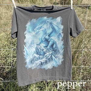 May include: A gray t-shirt featuring a detailed illustration of a blue dragon perched on a rocky, icy landscape. The dragon has large wings and is surrounded by ice formations. The word "pepper" is printed at the bottom.