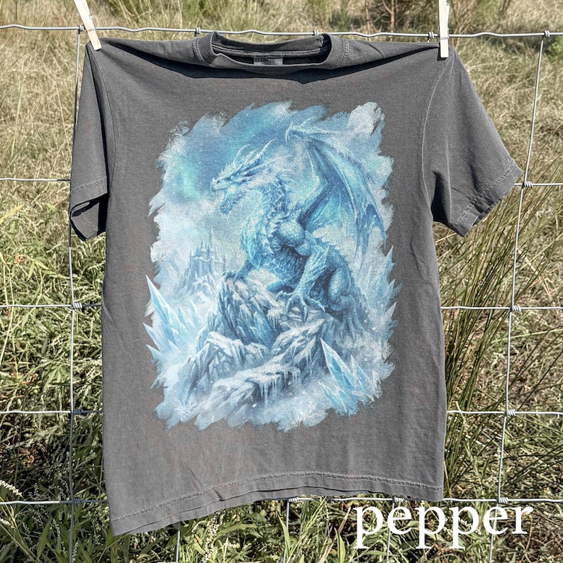 May include: A gray t-shirt featuring a detailed illustration of a blue dragon perched on a rocky, icy landscape. The dragon has large wings and is surrounded by ice formations. The word "pepper" is printed at the bottom.