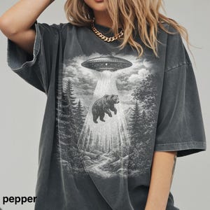 May include: A charcoal gray oversized t-shirt featuring a graphic print of a bear being abducted by a UFO in a forest setting. The design includes a beam of light shining down from the spacecraft onto the bear, surrounded by pine trees and clouds.