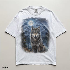 May include: White t-shirt featuring a detailed illustration of a wolf in a forest setting under a full moon. The wolf has gray and brown fur, with mountains and trees in the background. The text "TIMELESS COLORS" is visible on the upper right.