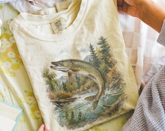 May include: Ivory-colored t-shirt featuring a detailed illustration of a fish leaping from a river, set against a backdrop of trees and mountains. The shirt is a Comfort Colors brand. The word "ivory" is printed at the bottom.
