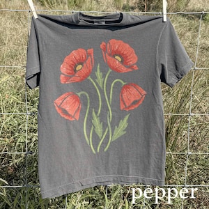 Op de afbeelding: Een grijs t-shirt met een bloemenontwerp van vier rode klaprozen met groene stelen en bladeren. Het woord "pepper" staat in witte letters onderaan. Het t-shirt hangt aan een waslijn.