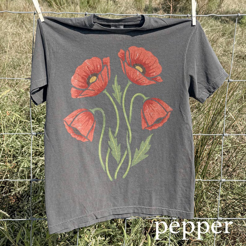 Op de afbeelding: Een grijs t-shirt met een bloemenontwerp van vier rode klaprozen met groene stelen en bladeren. Het woord "pepper" staat in witte letters onderaan. Het t-shirt hangt aan een waslijn.