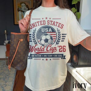 May include: An ivory t-shirt featuring a graphic print with the text UNITED STATES USA World Cup '26 ONE NATION ONE TEAM ONE GOAL. The design includes a soccer ball, a circular emblem with stars and stripes, and laurel leaf accents in red and blue.