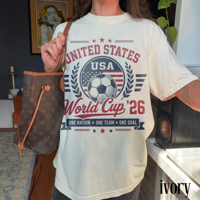 May include: An ivory t-shirt featuring a graphic print with the text UNITED STATES USA World Cup '26 ONE NATION ONE TEAM ONE GOAL. The design includes a soccer ball, a circular emblem with stars and stripes, and laurel leaf accents in red and blue.