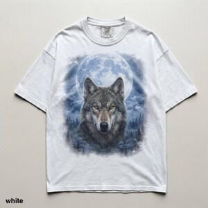 May include: White t-shirt featuring a detailed wolf portrait set against a full moon and forest backdrop. The wolf has gray fur and yellow eyes. The shirt has short sleeves and a crew neck. The text "white" is at the bottom of the shirt.
