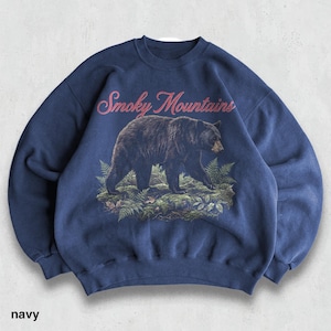 May include: Navy blue sweatshirt featuring a black bear walking through a forest scene. The words "Smoky Mountains" are arched across the top in pink script. The word "navy" is printed at the bottom of the sweatshirt.