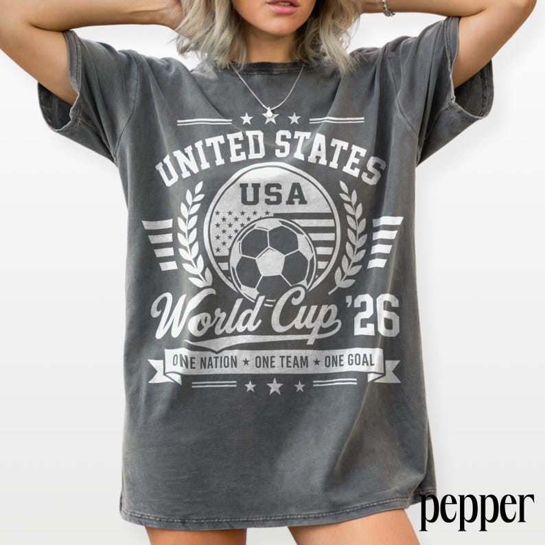 May include: A charcoal grey short-sleeve t-shirt featuring a white graphic print that reads UNITED STATES USA World Cup 26 ONE NATION ONE TEAM ONE GOAL with a central soccer ball icon surrounded by laurel leaves and stars in a vintage style.