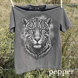 May include: A gray t-shirt featuring a detailed black and white illustration of a jaguar's face. The shirt is hanging on a clothesline. The word "pepper" is at the bottom of the image.