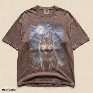 May include: Espresso-colored t-shirt featuring a detailed illustration of a wolf against a backdrop of a full moon, mountains, and trees. The wolf is the focal point, with a detailed fur pattern. The shirt has a classic crew neck and short sleeves.