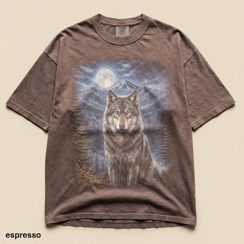 May include: Espresso-colored t-shirt featuring a detailed illustration of a wolf against a backdrop of a full moon, mountains, and trees. The wolf is the focal point, with a detailed fur pattern. The shirt has a classic crew neck and short sleeves.
