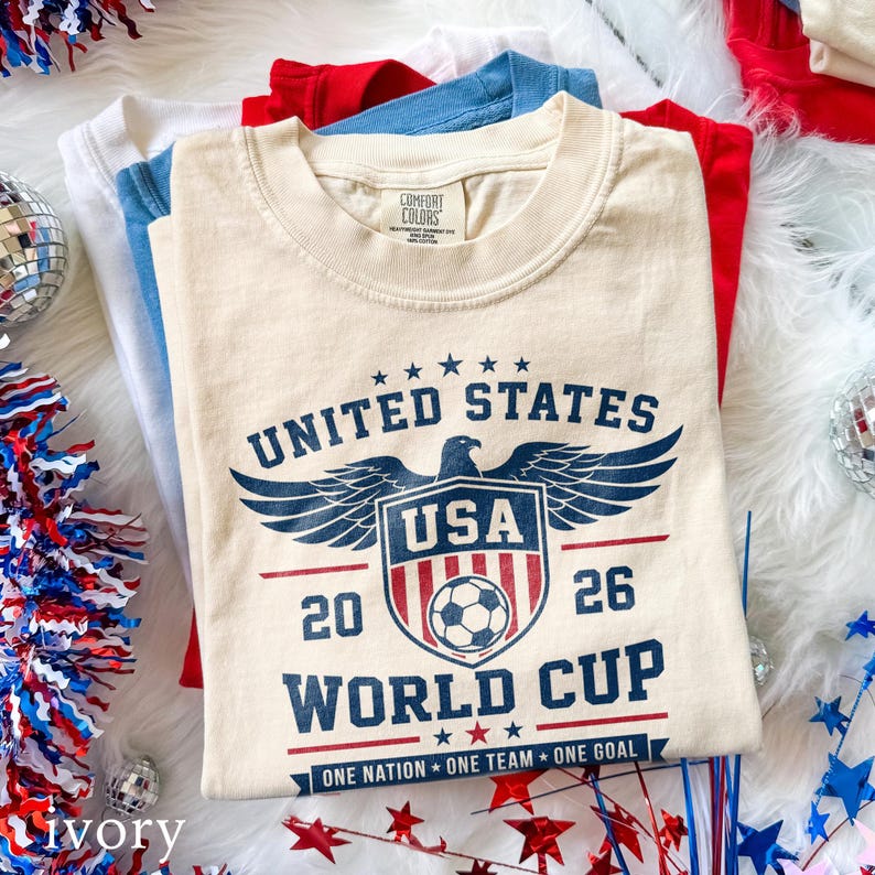 May include: An ivory crew neck t-shirt with a dark blue graphic print featuring an eagle, a shield with a soccer ball, and the text UNITED STATES 20 26 WORLD CUP ONE NATION ONE TEAM ONE GOAL. The shirt has a Comfort Colors brand label at the neck.