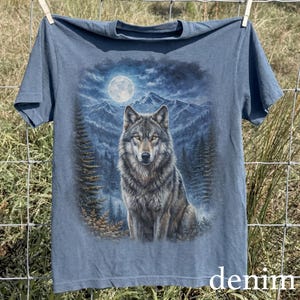 May include: Denim blue t-shirt featuring a detailed illustration of a wolf under a full moon in a forest setting. The wolf is the focal point, with mountains and trees in the background. The word "denim" is printed at the bottom.