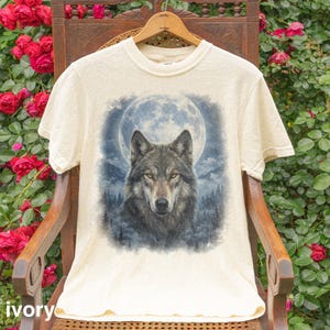 May include: Ivory-colored t-shirt featuring a detailed wolf portrait set against a full moon and forest backdrop. The wolf has gray and brown fur with yellow eyes. The word "ivory" is printed at the bottom.