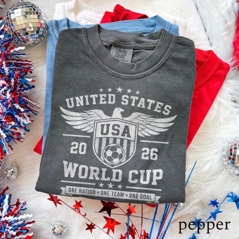May include: A charcoal gray short-sleeve t-shirt featuring a white graphic of an eagle above a shield with a soccer ball, the text UNITED STATES 20 26 WORLD CUP, and the phrase ONE NATION ONE TEAM ONE GOAL, with a Comfort Colors brand label.