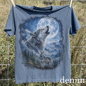May include: Denim blue t-shirt featuring a detailed graphic of two wolves howling at a full moon in a mountain landscape. The word "denim" is printed at the bottom. The shirt is hanging on a clothesline.
