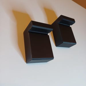 Op de afbeelding: Twee zwarte, haakse beugels met een rechthoekig ontwerp. De beugels zijn gemaakt van een solide materiaal en zijn op een wit oppervlak geplaatst. De beugels hebben een strakke, moderne uitstraling.