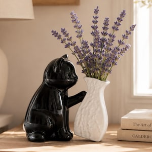 Pode incluir: Uma estatueta de gato em cerâmica preta brilhante sentada com uma pata apoiada contra um vaso branco texturizado. O vaso contém um buquê de ramos de lavanda roxos. O gato tem superfícies lisas e reflexivas e orelhas pontudas.